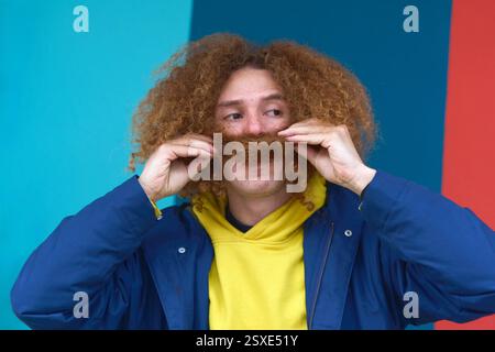 Persona dai capelli rossi con capelli ricci che tiene i baffi su uno sfondo colorato, indossa abiti casual e sorride Foto Stock