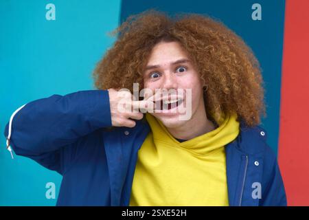 Giovane dai capelli ricci che tira giocosamente la bocca con un gesto umoristico su uno sfondo colorato, esprimendo gioia e spontaneità Foto Stock