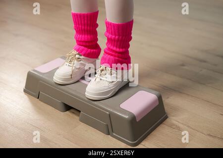 Vista ravvicinata dei piedi della persona con sneakers bianche e scaldasci rosa che si esercitano sulla piattaforma aerobica per passi in interni durante la sessione di allenamento Foto Stock