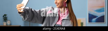 Donna con i capelli lunghi che sorride mentre scatta selfie in un ambiente moderno che indossa un abbigliamento casual dai colori pastello e accessori vivaci Foto Stock