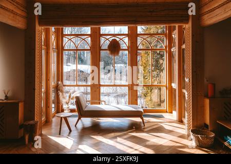 La luce del sole illumina gli intimi interni del tradizionale chalet in legno, caratterizzato da una confortevole chaise longue e grandi finestre che mostrano la scena invernale Foto Stock