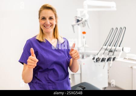 Ritratto del dentista felice nell'ufficio del dentista. Donna adulta sicura che lavora in una clinica odontoiatrica. Sta mostrando i pollici in alto. Foto Stock