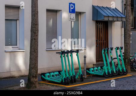 Una fila di scooter elettrici è parcheggiata ordinatamente in un'area designata accanto agli edifici residenziali. L'ambientazione è urbana, con alberi che gettano ombre su di esso Foto Stock