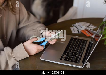 Giovane donna seduta a una scrivania di fronte al computer portatile e tagliando la carta di credito con le forbici Foto Stock