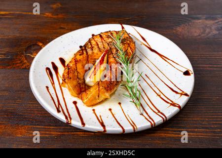 Un filetto di salmone perfettamente grigliato si trova su un piatto bianco, guarnito con un rametto di rosmarino e un filo di salsa. I colori ricchi e l'attenta preparazione Foto Stock