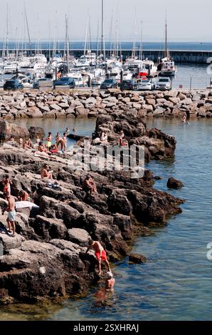 Bagnanti e nuotatori sulla costa rocciosa della spiaggia di Santa Marta, con un porticciolo pieno di barche a vela Foto Stock