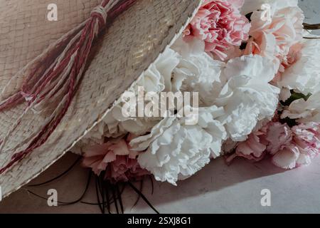 cappello di paglia estivo con doppio nastro rosa sopra un gruppo di teste di fiori di garofano rosa e bianco su un tavolo di marmo in stile vintage, spazio copia, angolo alto Foto Stock