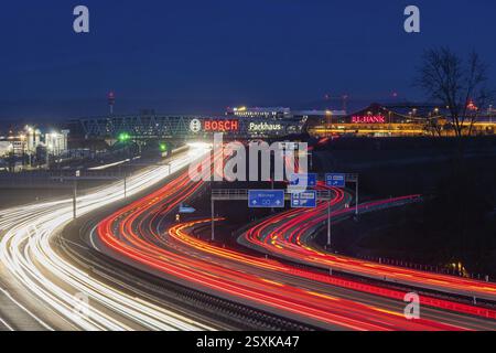 Autostrada A8 all'aeroporto di Stoccarda in serata. Traffico pesante, corsie di luce. Architettura moderna della Messe Stuttgart con parcheggio a più piani abov Foto Stock