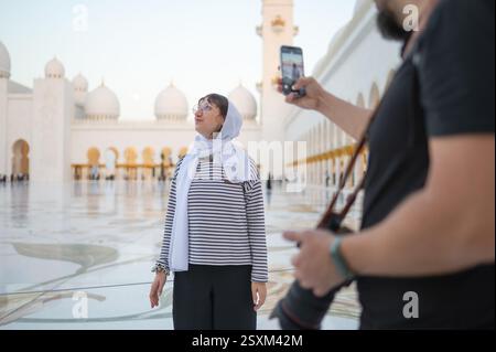 Un fotografo maschile scatta una foto di una donna con un foulard usando il suo telefono alla grande Moschea dello sceicco Zayed. Amici che visitano Abu Dhabi e creano Foto Stock