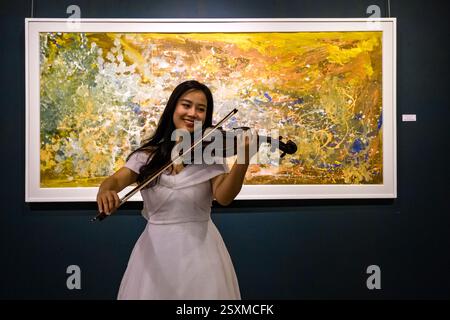 Una giovane donna sorridente con un abito bianco suona il violino davanti a un dipinto in stile astratto. Foto Stock