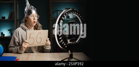 Una strana blogger con cappello in lamina che mostra un messaggio 5G che parla con i suoi iscritti sulla fotocamera, registra in diretta streaming a casa. Notizie false e cospirazione Foto Stock