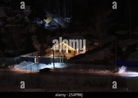 Luci dorate soffuse creano un'atmosfera accogliente in una tranquilla notte d'inverno. La neve ricopre il terreno mentre le case lontane brillano dolcemente, evocando la pace Foto Stock