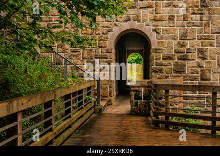 Un arco di pietra splendidamente strutturato incornicia una vista panoramica oltre, che conduce attraverso una passerella rustica in legno circondata dal verde. Foto Stock
