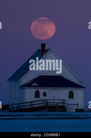Grande luna piena che sorge su una piccola chiesa bianca in un paesaggio innevato Foto Stock