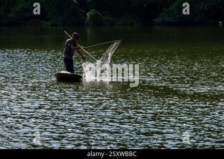 Un pescatore tradizionalmente pesca con una rete da una piccola barca su un fiume. Foto Stock