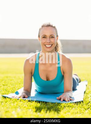 Donna sorridente che pratica yoga sul tappeto all'aperto, godendosi la mattina di sole Foto Stock