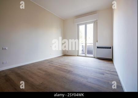 Appartamento vuoto con pavimento in legno, pareti bianche, porta del balcone e radiatore, ideale per lavori di ristrutturazione o progetti di interior design Foto Stock