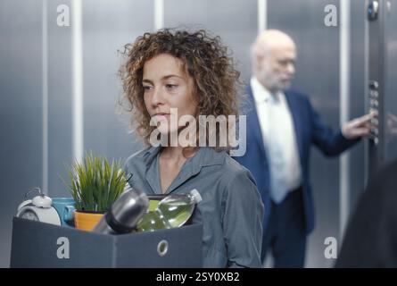 Donna d'affari che porta una scatola con i suoi effetti personali e scende dall'ascensore, ha perso il lavoro Foto Stock