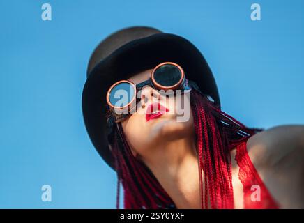 Una ragazza glamour sfoggia i suoi dreadlock scarlatti indossando un costume da bagno rosso, un cappello nero e occhiali da saldatura sotto il cielo blu brillante. La scena Foto Stock