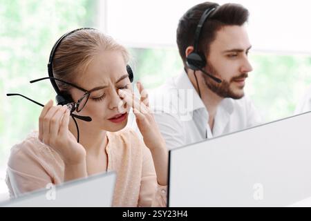 Operatore hotline sovraccarico di lavoro davanti al monitor del computer e i suoi colleghi impegnati presso l'ufficio del call center Foto Stock