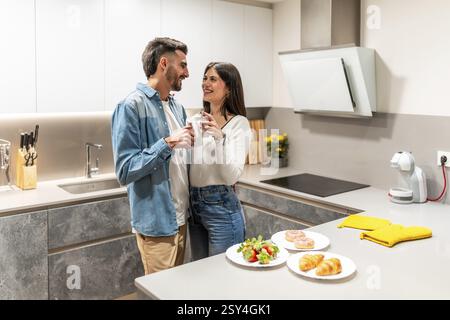 Giovani coppie che abbracciano e bevono caffè nella loro cucina moderna, con pasticcini per la colazione e frutta fresca sul bancone Foto Stock