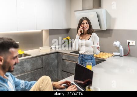 Giovane donna che chatta al telefono mentre si gusta una tazza, mentre l'uomo lavora su un notebook in una cucina moderna e luminosa, creando un'atmosfera accogliente Foto Stock