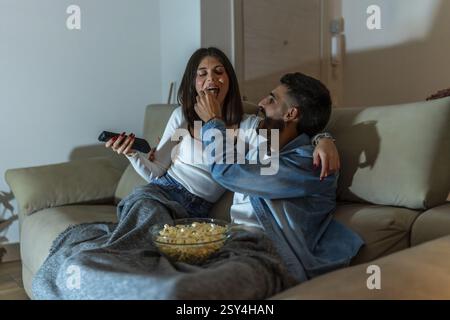 Giovani coppie che si rilassano sul divano, gustando popcorn e guardando la televisione insieme durante una notte intima nel loro confortevole soggiorno Foto Stock