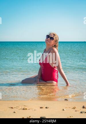 Una donna con un costume da bagno rosso siede sulla spiaggia sabbiosa con le onde che le lambiscono dolcemente. Foto Stock