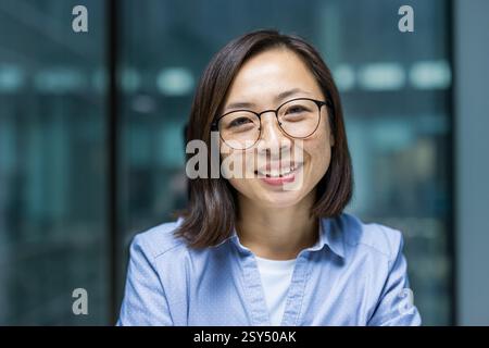 Primo piano di una donna d'affari asiatica sorridente con gli occhiali, che indossa una camicia azzurra in un moderno ambiente da ufficio. Foto Stock