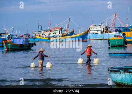 Due donne con cappelli conici trasportano acqua in barattoli nell'acqua di fronte alla spiaggia di Mui ne, Mũi ne, molte barche da pesca in lontananza. Foto Stock