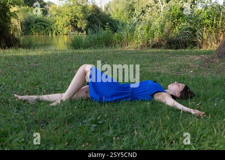 Donna che indossa un abito blu distesa sull'erba e si rilassa sul fiume in una giornata estiva di sole Foto Stock