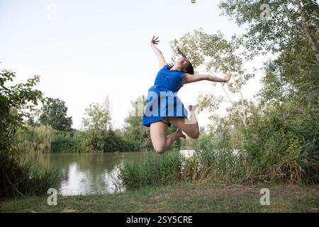 Giovane donna spensierata che indossa un abito blu che salta vicino al lago, godendo della libertà e della natura Foto Stock