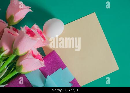 Composizione piatta con rose rosa, regalo avvolto, uovo e carta bianca su sfondo verde colorato. Foto Stock