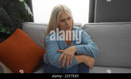 Donna seduta su un divano in un soggiorno con un'espressione premurosa che indossa un maglione blu circondato da un accogliente arredamento di casa, tra cui un cuscino arancione Foto Stock