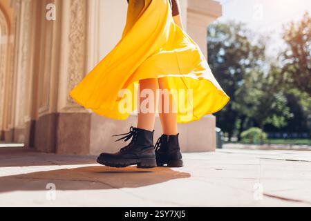 Donna che indossa un outfit con gonna gialla e stivali in pelle nera in città. Primo piano di scarpe e tessuto fluido in movimento. Vestiti femminili. Foto Stock