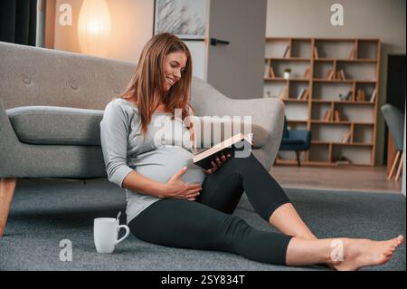 Seduto sul pavimento e leggendo il libro. Una bella donna incinta è al chiuso a casa. Foto Stock