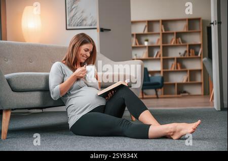 Seduto sul pavimento e leggendo il libro. Una bella donna incinta è al chiuso a casa. Foto Stock