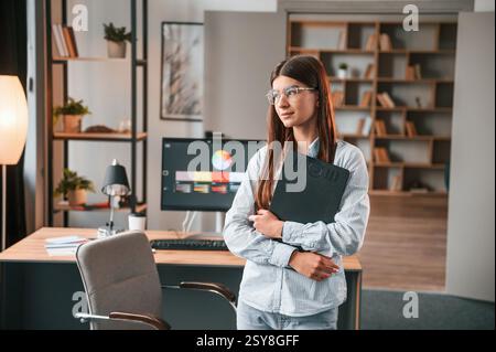 Con blocco note nero. In piedi e in posa. Una giovane designer donna è nell'ufficio moderno. Foto Stock