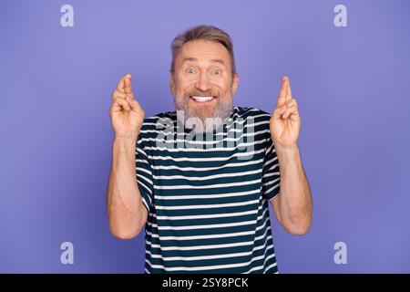 Uomo anziano allegro sorridente con le dita incrociate davanti a uno sfondo viola e indossa un'elegante T-shirt a righe Foto Stock