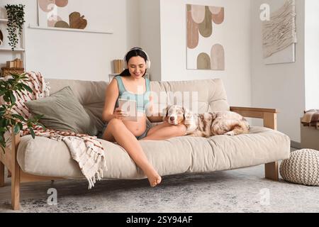 Giovane donna incinta sportiva in cuffie con tablet e simpatico cane pastore australiano seduto sul divano di casa Foto Stock