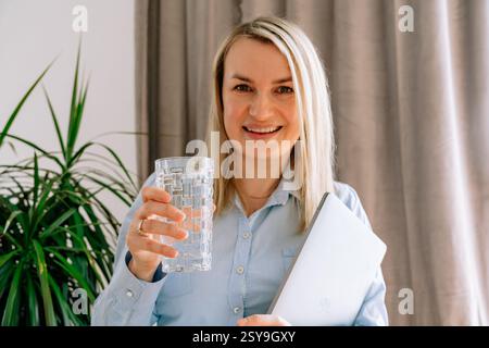 Ragazza sorridente che tiene un bicchiere d'acqua seduta a tavola, 35-37 anni di aspetto europeo da vicino vestito con una camicia blu di luce naturale Foto Stock