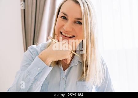 Ragazza sorridente che tiene le mani vicino al viso tenendo una penna, 35-37 anni di aspetto europeo da vicino vestito con una camicia blu. Foto di alta qualità Foto Stock