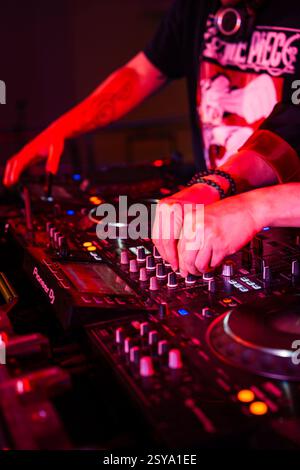 Il DJ mette le mani su un mixer, due DJ suonano sul tabellone e il club è illuminato da luci rosse Foto Stock