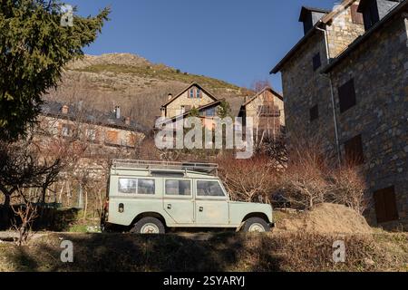 Un vecchio e classico veicolo fuoristrada parcheggiato di fronte alle tradizionali case in pietra in un pittoresco villaggio rurale di montagna, circondato da alberi aridi e colline Foto Stock