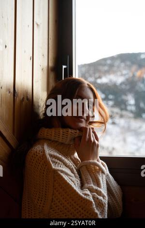Una giovane donna con i capelli rossi gode di un'accogliente giornata invernale all'interno, vestita calorosamente con un maglione a maglia, sorridendo dolcemente mentre guarda fuori dalla finestra. Foto Stock
