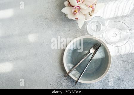 Vista dall'alto di un ambiente da pranzo minimalista. Una piastra grigia incrinata con forchetta e coltello, completata da bicchieri di cristallo e un'orchidea bianca su un textur Foto Stock