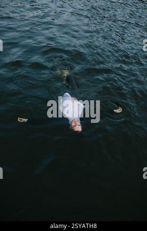 Un uomo vestito con una tuta galleggia sull'acqua scura con le braccia allungate, creando una scena surreale e introspettiva. L'acqua increspata e l'abbigliamento formale Foto Stock