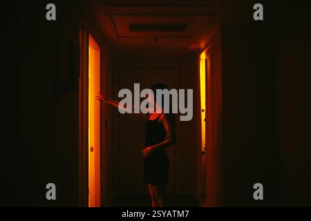 Silhouette di una giovane donna in un abito scuro che apre una porta luminosa della camera d'albergo, evidenziando l'intrigante contrasto tra l'oscurità e la luce Foto Stock