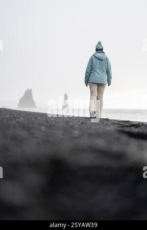 Vista posteriore di una donna solitaria irriconoscibile si dirige verso le famose faraglioni di mare di Reynisdrangar sulle nebbiose sabbie nere di Reynisfjara Beach i. Foto Stock