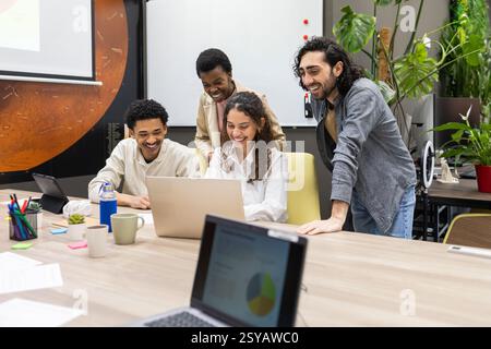 Un team diversificato di professionisti riuniti intorno a un notebook, collaborando con entusiasmo in un ufficio moderno. L'impostazione è vivace e piena di gr Foto Stock